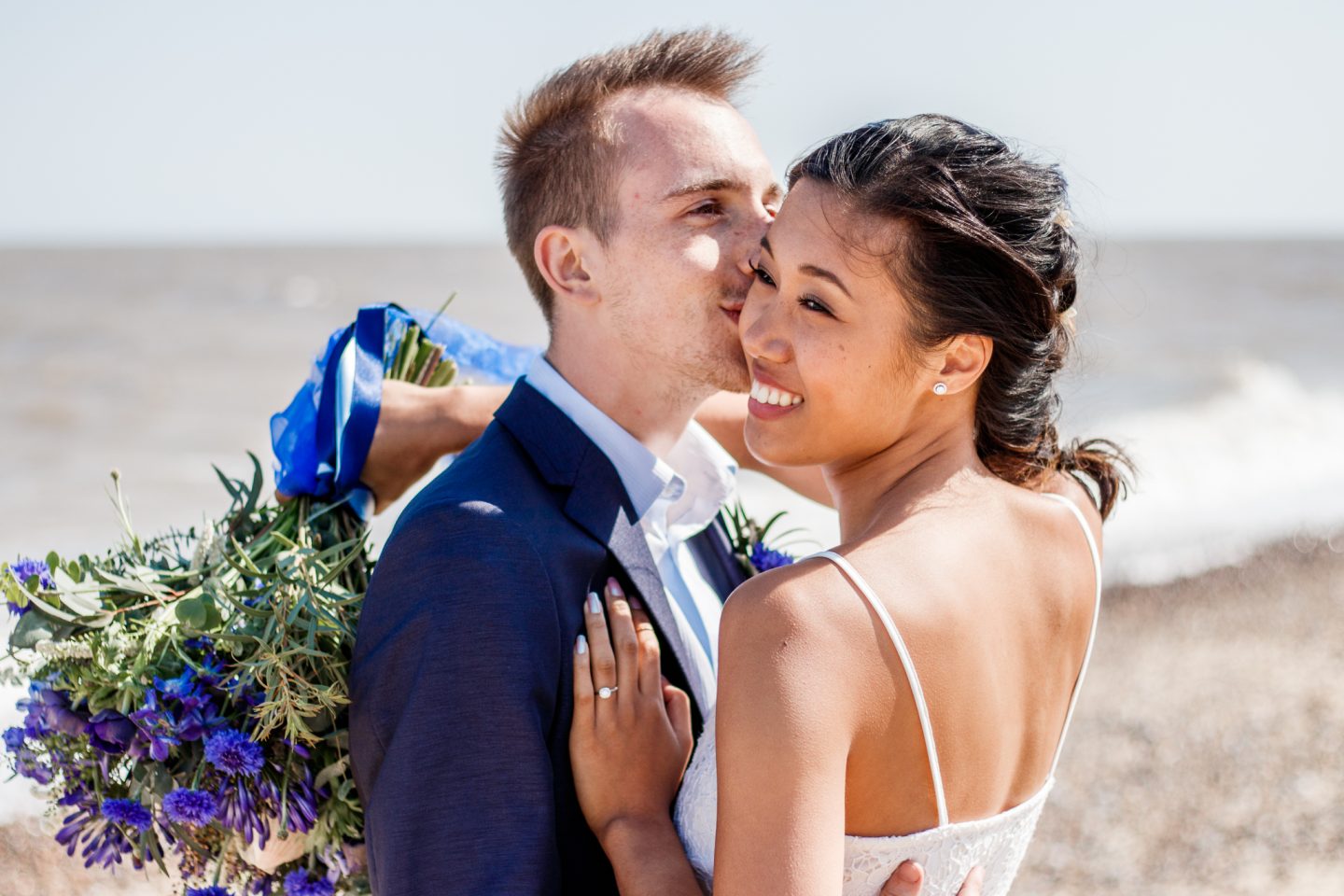 A wedding couple embracing on the beach wearing blue wedding dress holding a bouquet.